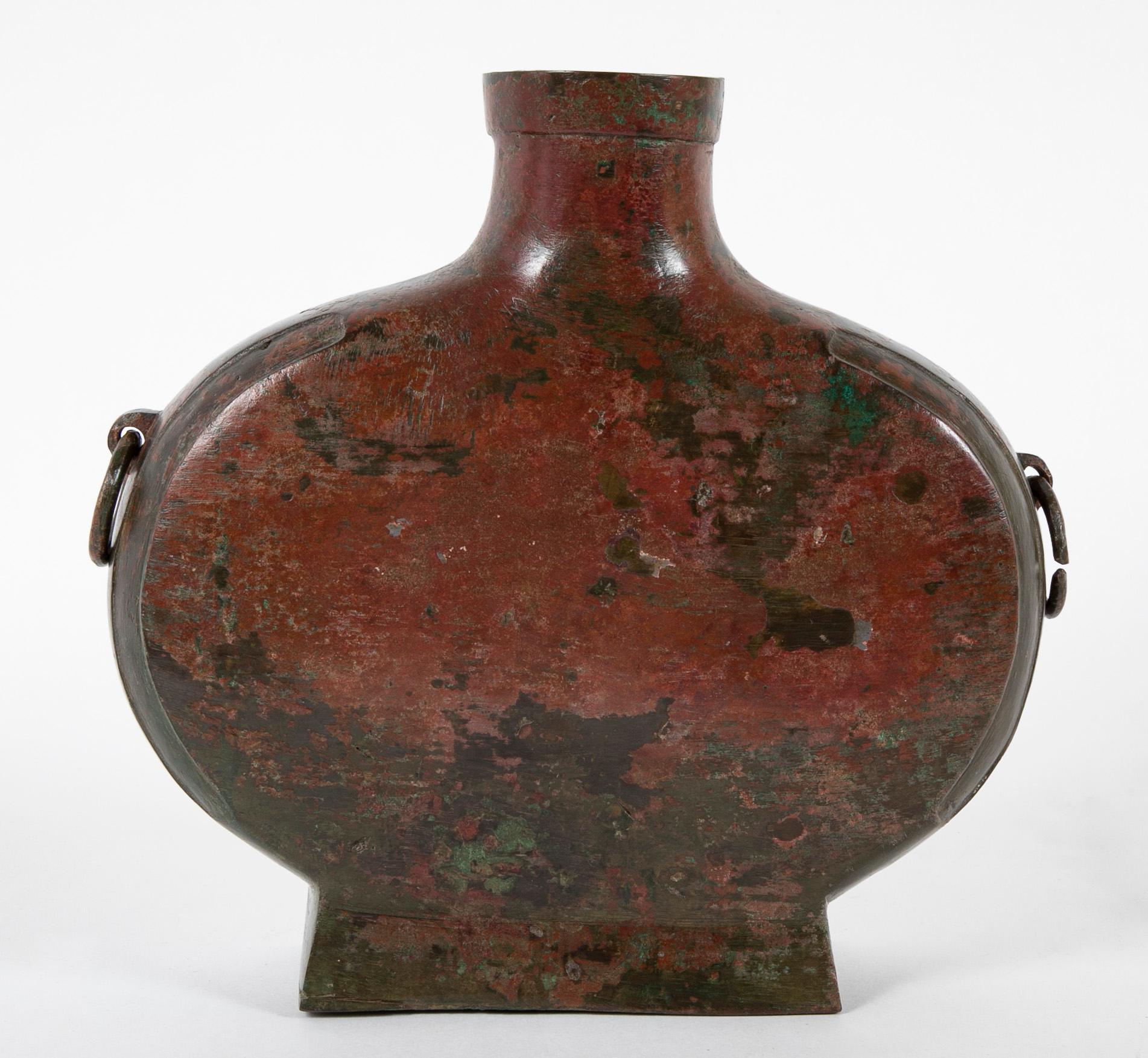 An archaic bronze vessel in flattened flask form with ring handles and mottled red and green patination.