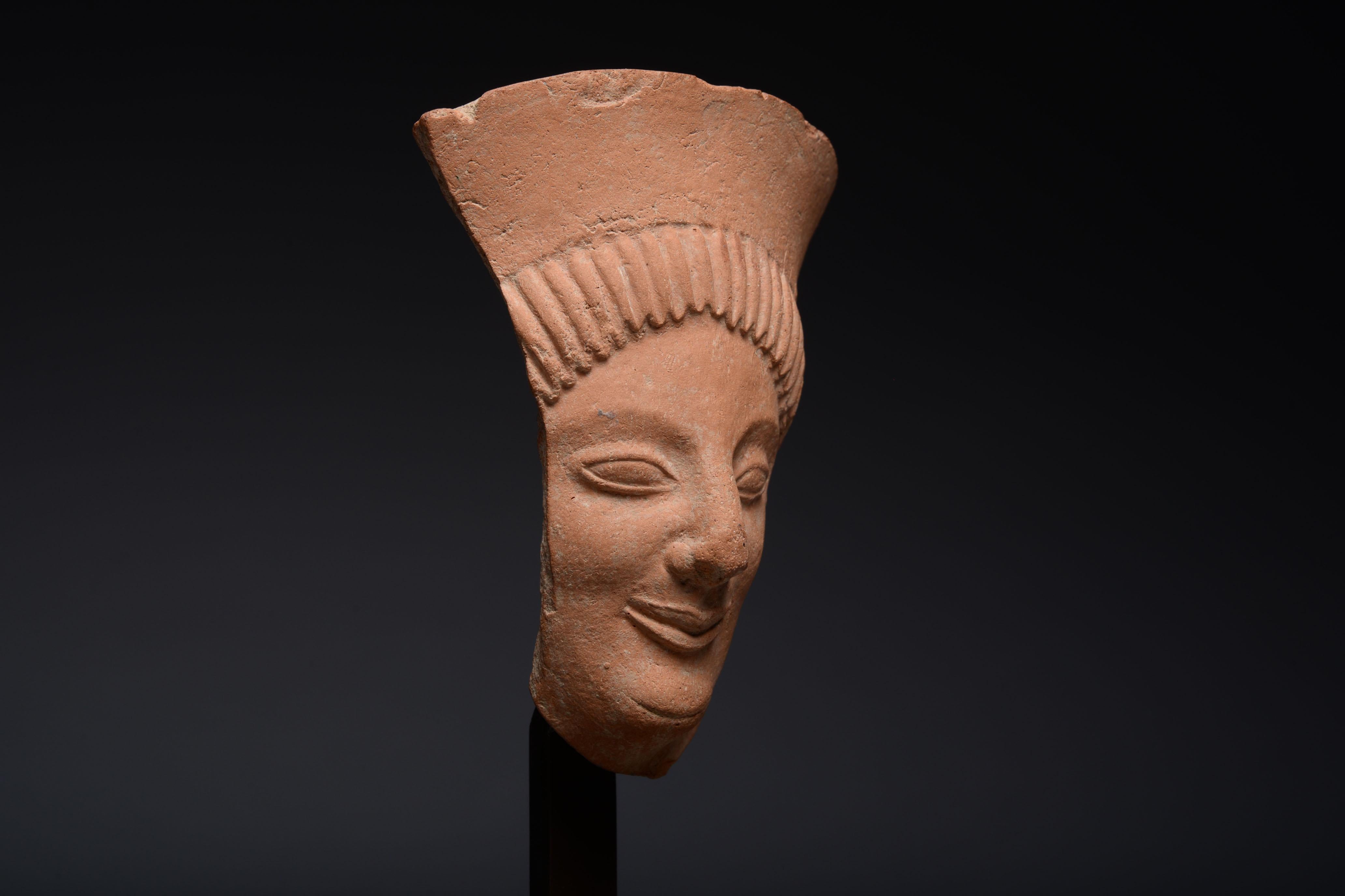 Greek terracotta protome fragment, archaic period, circa 6th century BC.

Depicting the face of a goddess with almond-shaped eyes, archaic smile and rounded chin, her hair gathered into neatly arranged pleats and covered with a tall