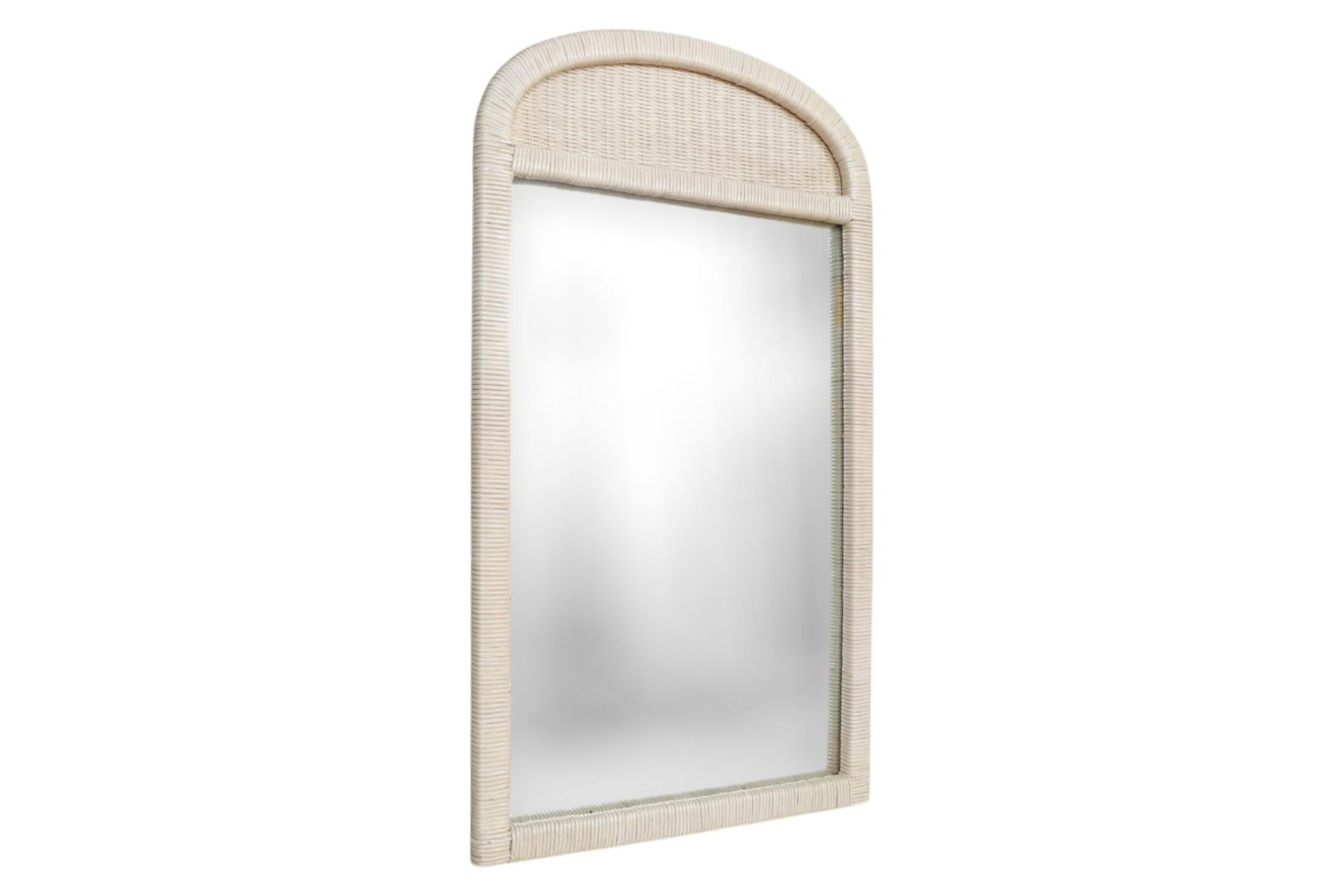 A white-washed coastal wall mirror attributed to Henry Link. The vertical wooden frame is wrapped with rattan, surmounted with a wicker arch. Manufacturer's mark in the back reads ‘Made in USA’.