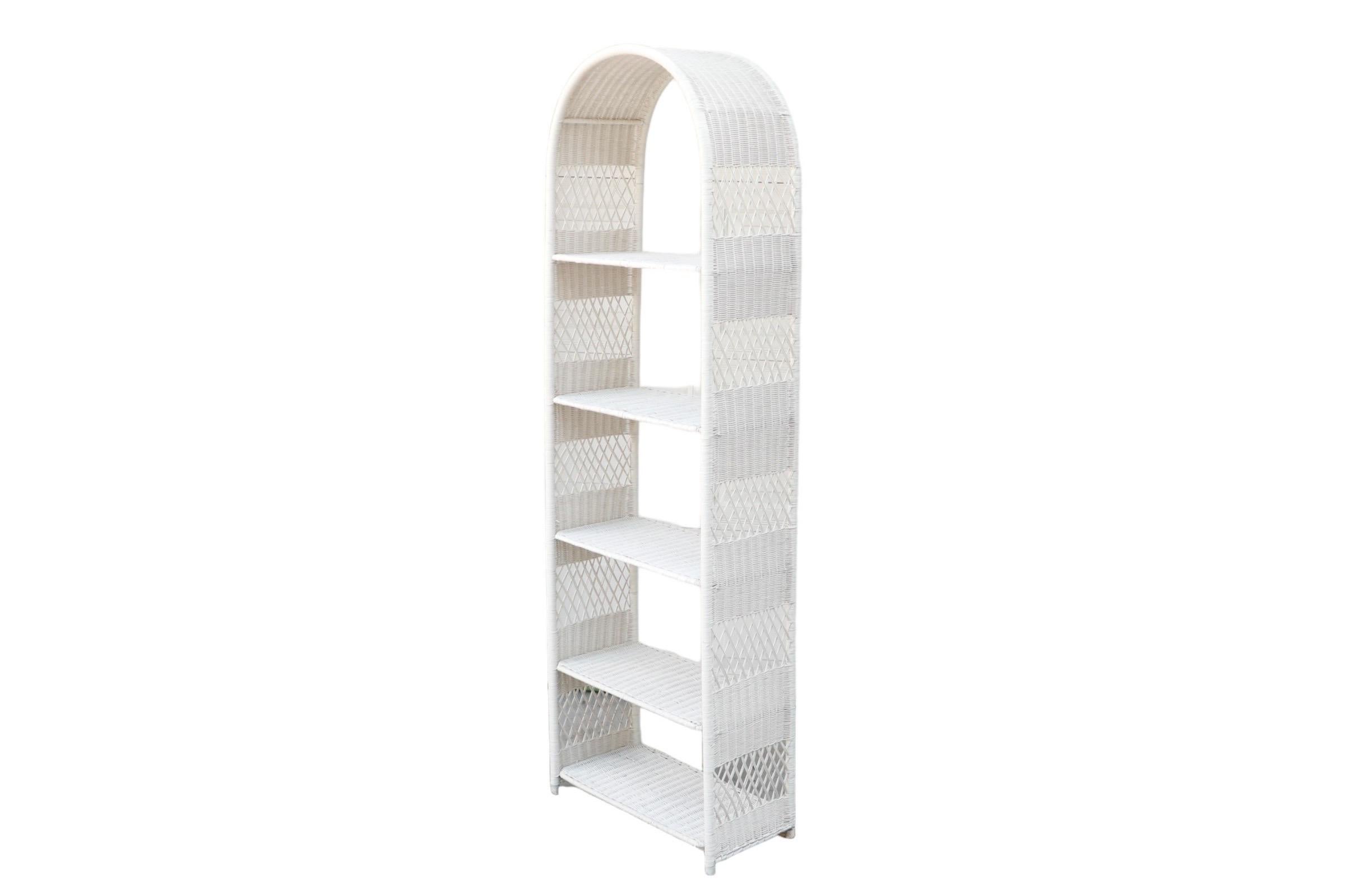 An open wicker etagere or bookcase in white, made of woven rattan. Five rattan shelves are set in an arched frame, with woven rattan and braided lattice panels at each side.