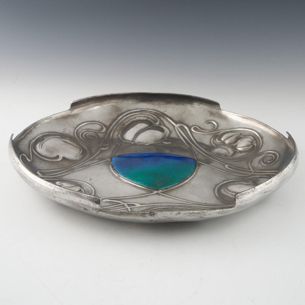 Heading : Archibald Knox for Liberty & Co polished pewter and enamelled dish 
Date : c1902-1905
Period : Edward VII 
Origin : England 
Decoration : Art Nouveau floral repousse decoration and turquoise and blue silver backed enamelled centre. 
Size :