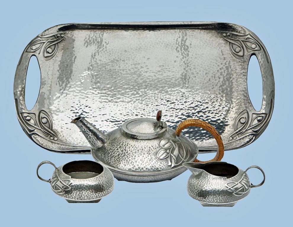 Archibald Knox Liberty Tudric hammered pewter tea set, circa 1906. Teapot, cream jug and sugar, each of compressed oval form with relief whiplash curved Celtic ornament interlace decoration, original wicker handle and button to teapot. Stamped six