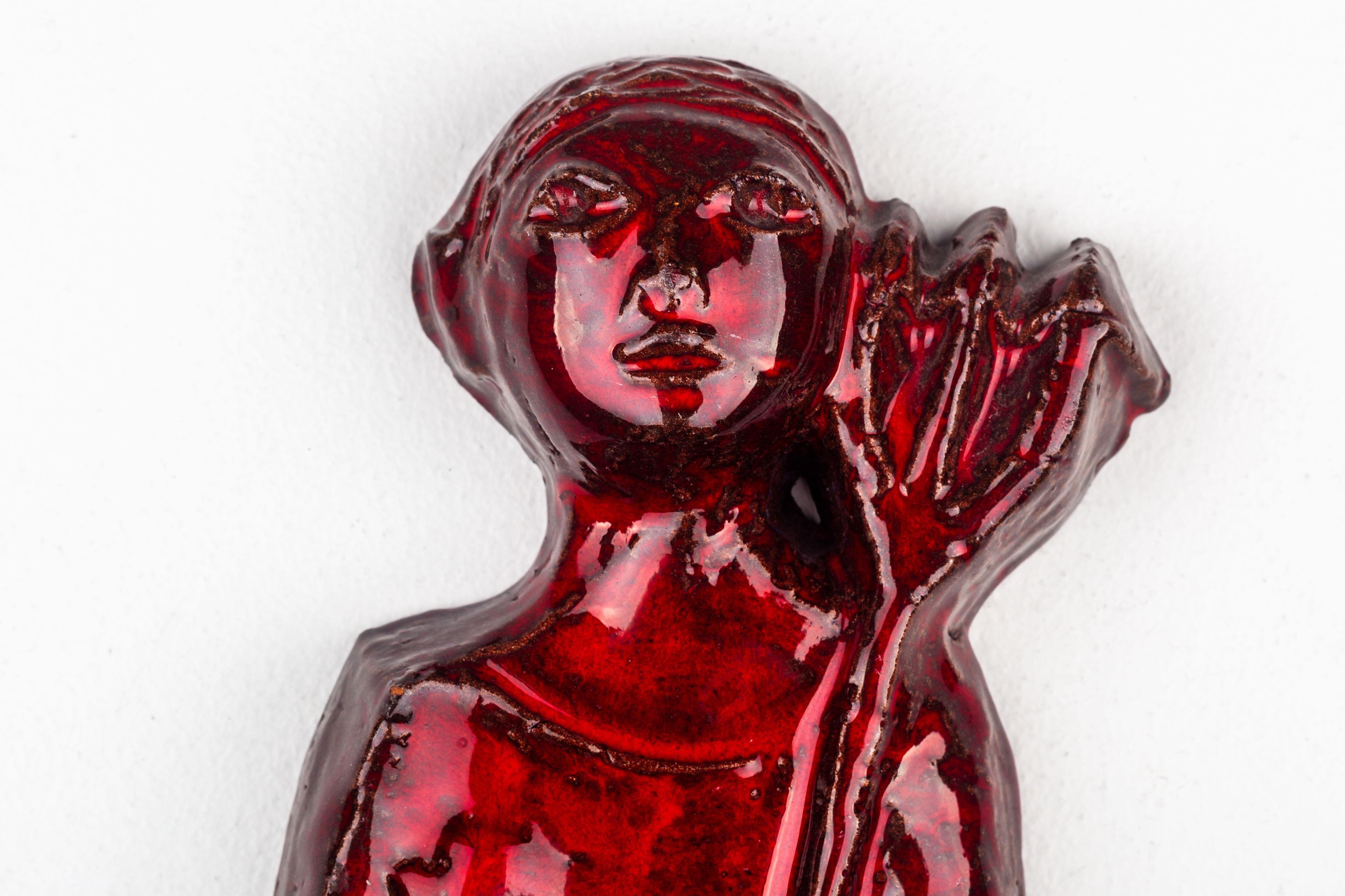 This ceramic wall decoration, a product of modernist Studio Pottery in Europe, portrays an architectural red devil holding a pitchfork amidst flames. The piece is characterized by vibrant red enamel tones, which shine through a super glossy clear