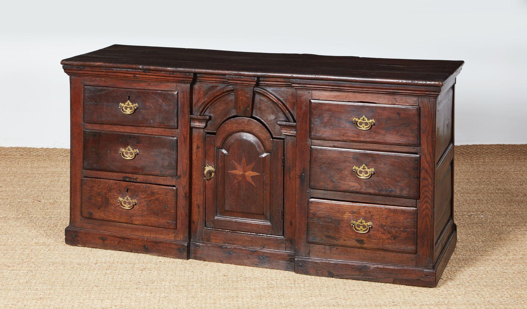 Comò basso in quercia gallese del XVIII secolo dalla forte forma architettonica, con un piano semplice a listelli e due banchi di cassetti che fiancheggiano un'anta centrale ad arco con chiave di volta e intarsio di stelle a compasso, e poggiante su