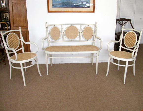 The two seater salon and chairs have; caned seats and backs, arm rests, some turning to uprights ending with finials, under stretchers, and slightly splayed legs. Set is thought to be by J Kohn. This set has been used in several Hollywood films,