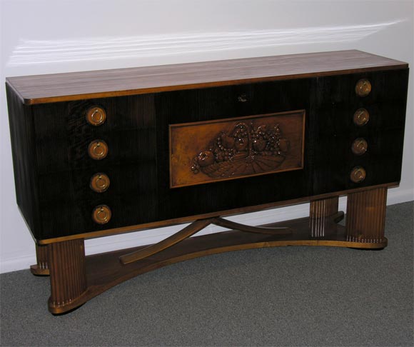 An Art Deco sideboard featuring eight drawers, a interior bar cabinet with drop front central door with a carved relief of a basket of fruit and oval shaped reeded legs on a shaped platform with a centered decorative 
