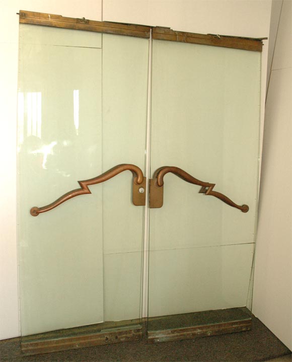 Impressive pair of original Art Deco doors with unique left and right facing bronze stylized scroll handles on both sides of each clear glass panel, framed at top and bottom by matching patinated bronze bands.  (Could even be fitted as great