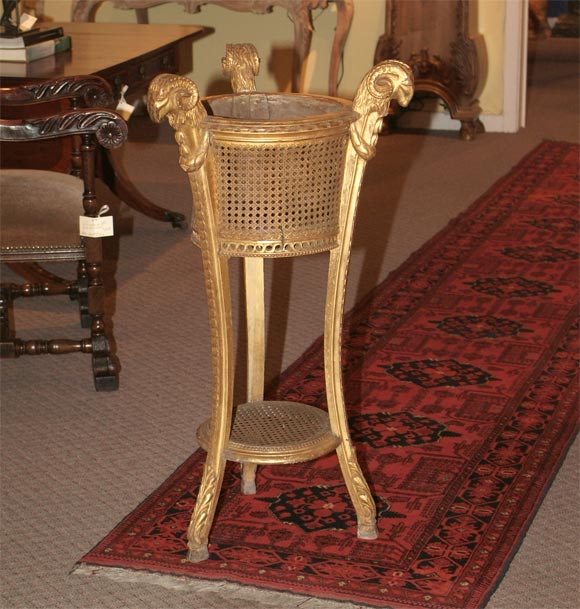 A carved and painted giltwood and cane jardiniere with the central round and second shelf supported by three ram's head topped acanthus carved legs.