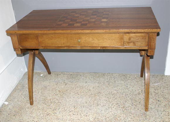 Richly figured walnut forms the body of this table, which is centered with an inlaid chess board in light and dark fruitwoods and supported on scissored sabre legs. A large plinth drawer on one side is lined in original (worn) florentine bookpapers.