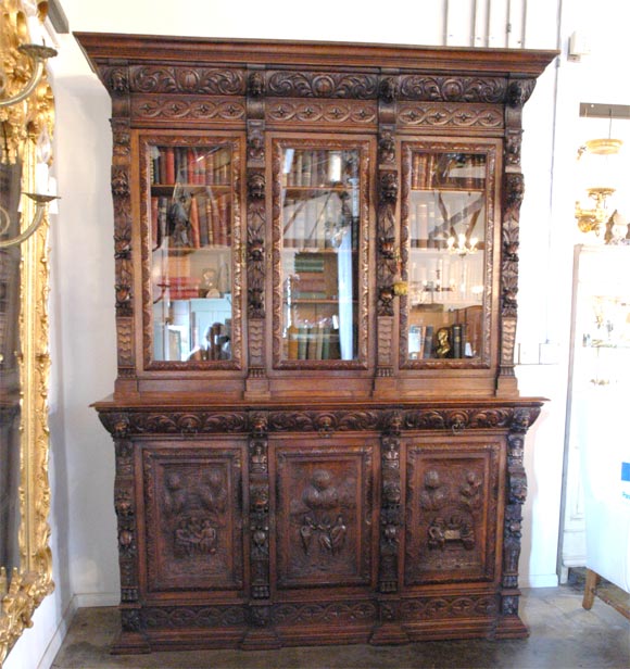 This bookcase has three glazed upper doors over three drawers and then three carved panel lower doors. The lower doors are highly carved and show various rural settings with people. There are many carved elements of: flowers, fruit, leaves, 1/2