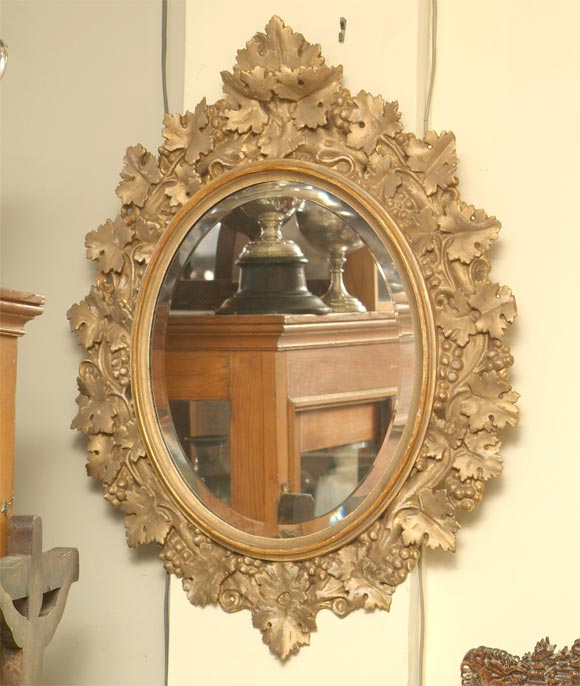 Specchio ovale in legno intagliato a mano con cornice in vetro smussato, con intagli di uva e foglie d'uva e finitura in vernice dorata.
Americano, circa 1880.
 