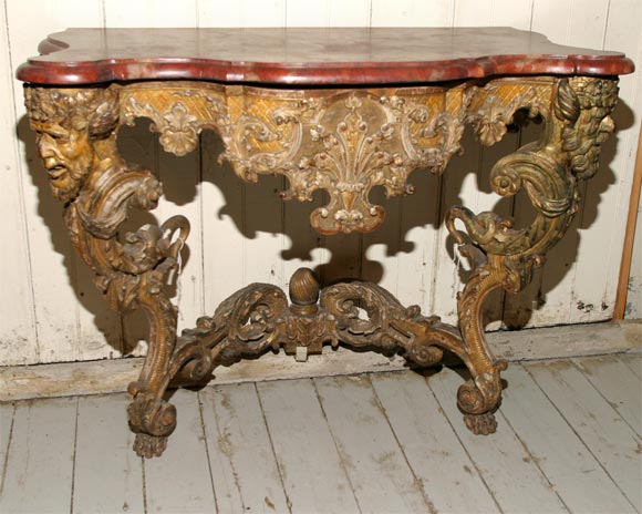 A German Baroque console with associated shaped faux marble top supported by satyrs' masks above bold foliate scroll legs, flanking an apron carved with rocaille decoration, the in-curved stretcher carved on both sides, with paw feet. The design of