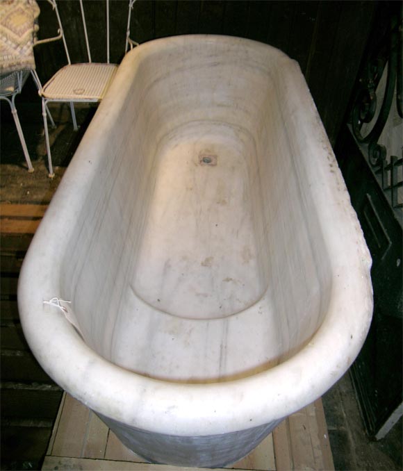 White carrera marble oval form bathtub with three smooth sides. Front side is decorated with two carved rings. Rear, long side is rough and meant to face a wall. The interior is formed with a slightly elevated area that serves as a seat.