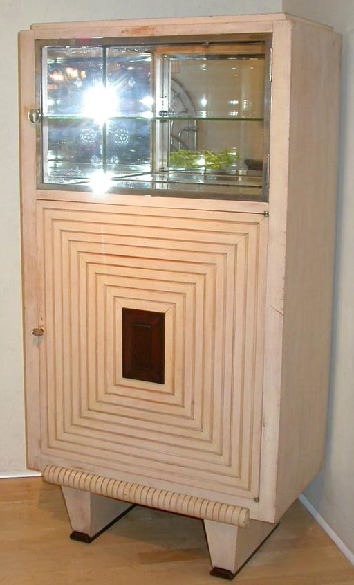 An Art Deco period cocktail cabinet having a painted finish with panels (including the top surface) in natural mahogany, the chrome plated metal framed upper door enclosing one glass shelf within a mirrored compartment, the lower door revealing