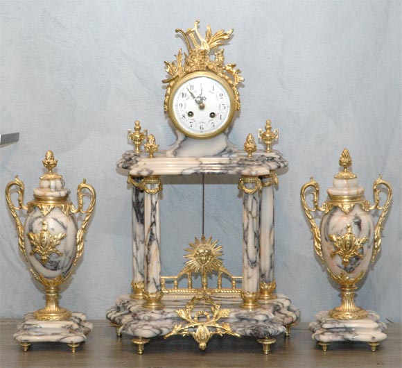 A French Louis XV-late 19th century gilt-bronze and marble mantle clock set. The hand painted enamel dial surmounted with a lyre. The eight day movement strikes on a bell. A pair of closed urns complete the ensemble. Jefferson West Antiques offer a