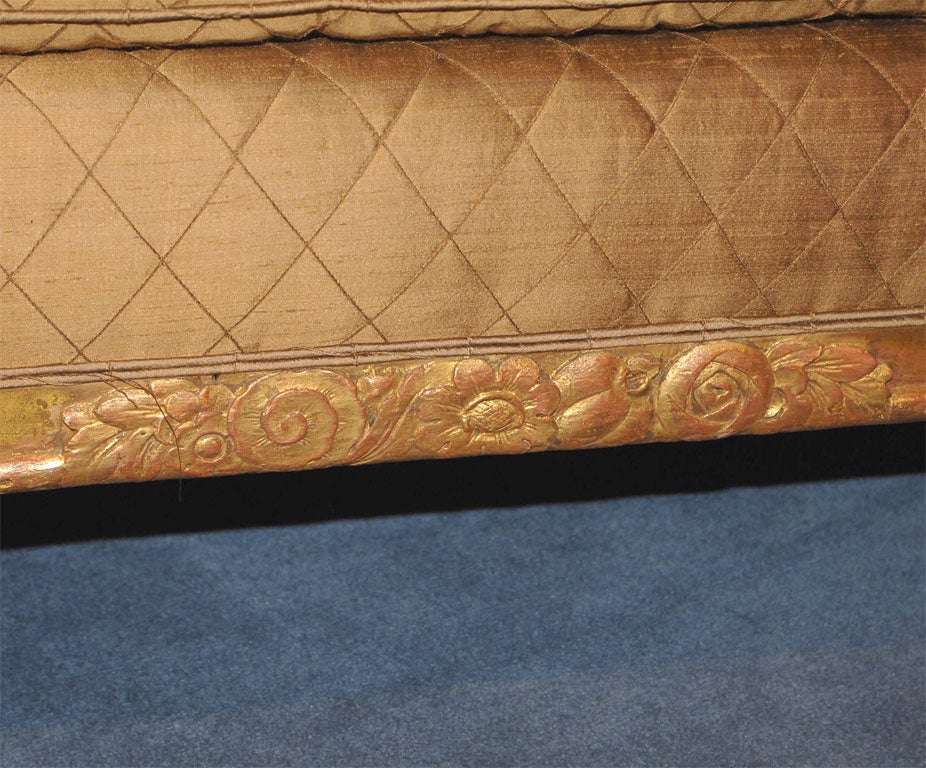 With arched sides carved with flowering foliage flanking a rectangular cushion above a carved rail, raised on dramatically tapering reeded legs; newly upholstered in bronze colored silk with a trelliswork design.