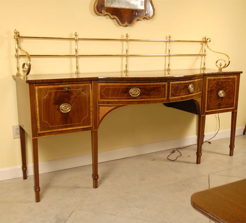 A very large (more than 7' wide) inlaid sideboard of Hepplewhite-inspired design, with a bow-front central section and resting on 6 square-tapered legs ending in 'block' feet.<br />
<br />
Resting on the serving platform is a brass 