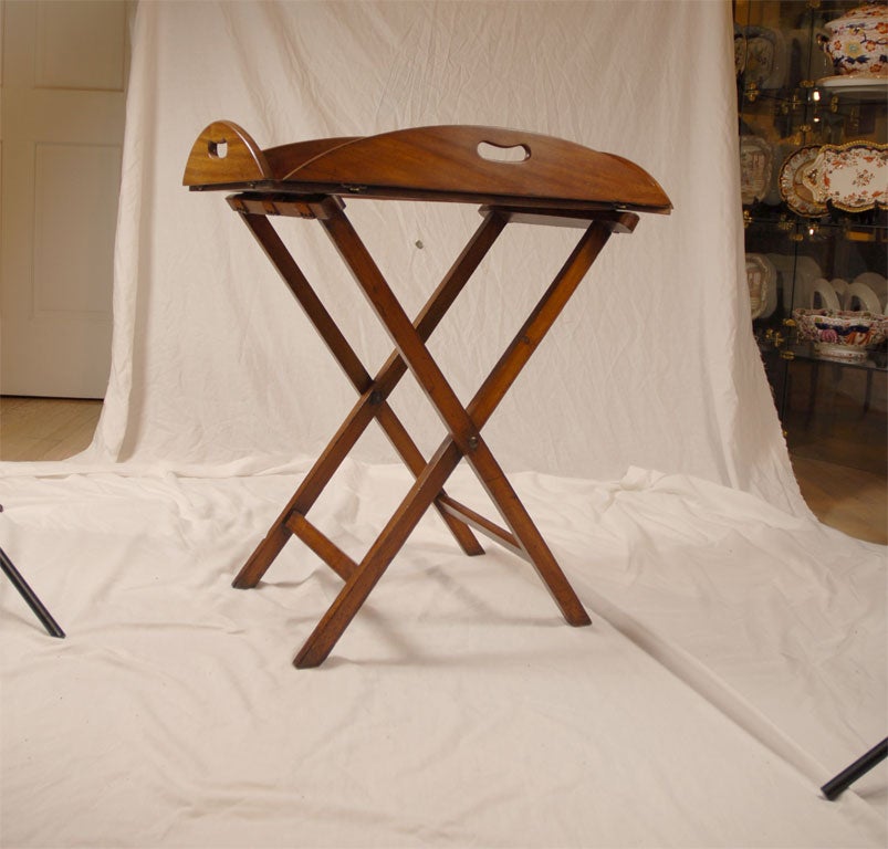 A mahogany butler's tray on stand, with an oval form that converts via metal hinges to a rectangle for carrying, by four kidney-shaped handles. Resting on a cross-braced stand connected by bolts and three fabric straps.<br />
<br />
c. 1810 England