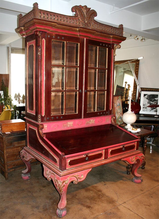 A large secretary in a Dutch Colonial style in red with gold detail, with double paned glass doors and desk top that slides open.