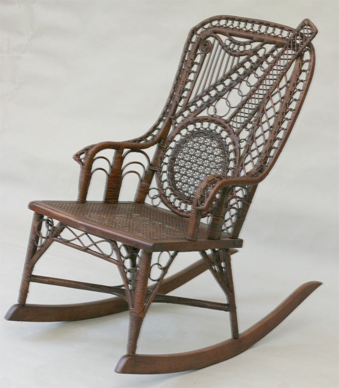 Ornate Victorian motif wicker rocking chair depicting two musical instruments on the back panel:  a banjo and a harp woven from reeds and strands of cane.  Intricate patterns and looping.  Pressed cane seat.