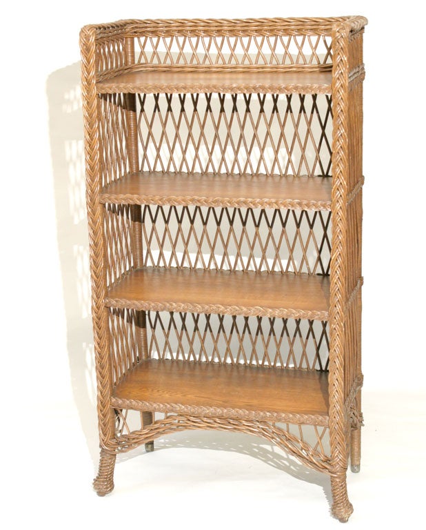 Bar Harbor wicker bookcase in natural stained finish.  Four solid oak shelves bordered in braidwork.  Three-sided wicker gallery on top.  Traditional open crisscrossed latticing overall.  Pineapple front feet, brass-capped rear feet.  Arched wicker