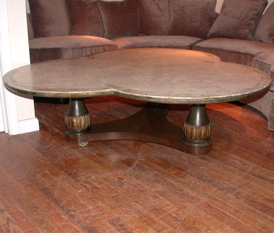 unusual large clover shaped travetine marble coffee table supported by a trio of detailed gilt leafed columns that are on a triangular base with casters for easy movement.