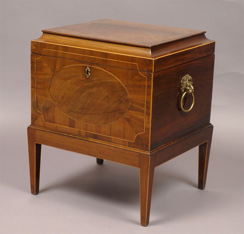 A late George III mahogany cellarette on stand. Constructed of mahogany, with a fine flame-mahogany veneer and stringing inlay. Mounted on either side with brass ring pulls clutched by a lion's mask mount. <br />
<br />
Top mounted with brass