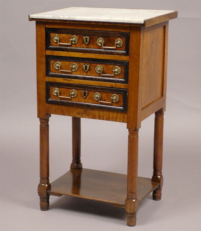 A three drawer bedside table in Walnut with a white marble top and turned legs. The marble top is inset over three shallow drawers, each mounted with brass keyhole escutcheons and handles. 