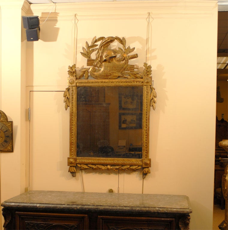 Miroir en bois doré de la fin de la période Louis XVI. La plaque de miroir est bordée d'un cadre néoclassique en bois doré avec des motifs de paterae sculptés. La partie supérieure est ornée d'un trophée en bois doré sculpté avec des symboles de la