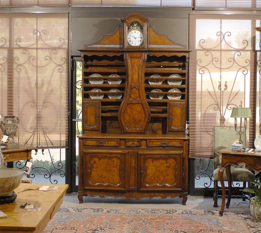 A fine French burled wood Vaisellier, mounted with a longcase clock in the center, and with deep carved designs accenting the walnut body. The body composed of an upper section with open plate racks and two hinged doors, above the lower buffet