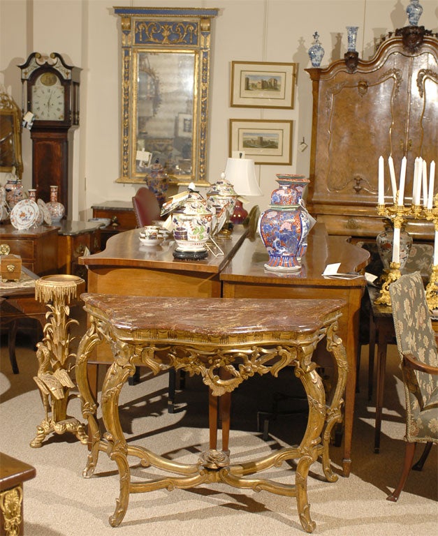 An Italian Gilt Wood console, mounted with a faux-marble top over scrolled legs. The gilt-wood legs braced with a crossed stretcher, at the intersection mounted with a rosebud finial. The top a wooden surface with applied painted material in maroon