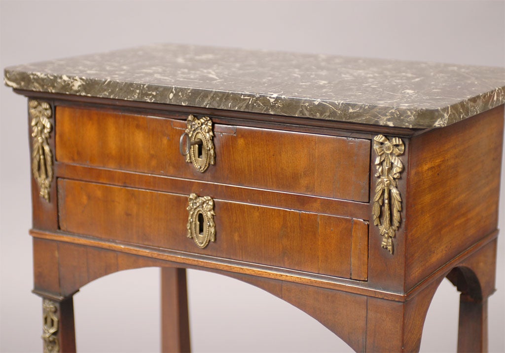 A Chiffonniere/bedside table in walnut, with fixtures in gilt-bronze and a deep grey and white marble table surface. French in origin, dating from the 19th century.

The table with two sliding drawers fixed with keyhole escutcheons in gilt-bronze