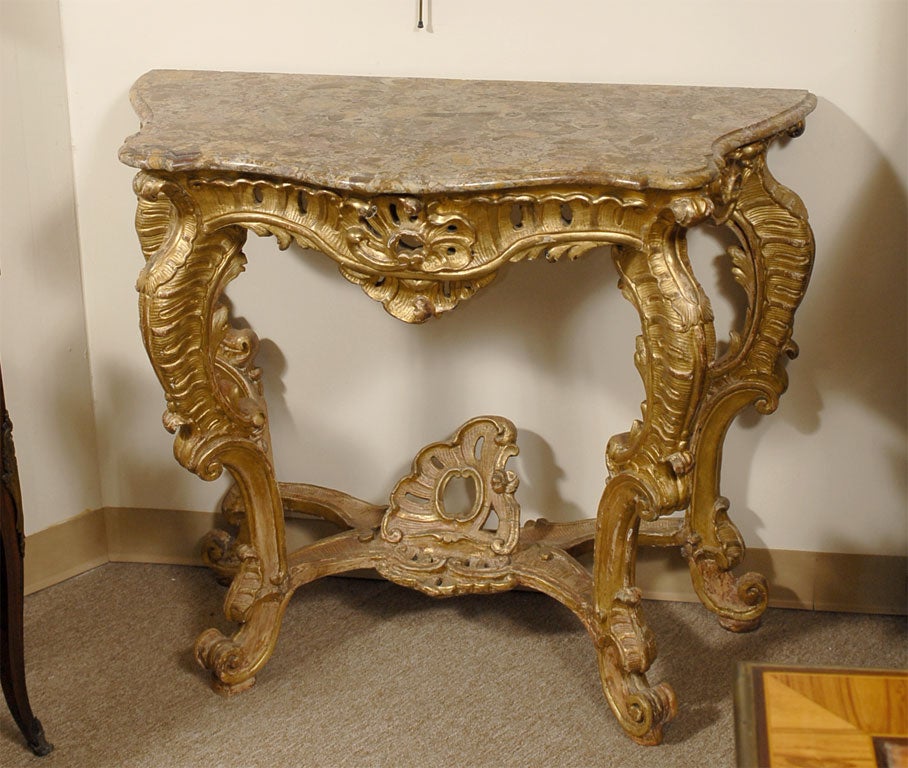 The Rococo console table with marble serpentine top on a pierced apron, featuring deep carved designs, all mounted on four cabriole legs ending with scroll feet. The base connected with a crossed stretcher, centered by a raised finial in cabochon