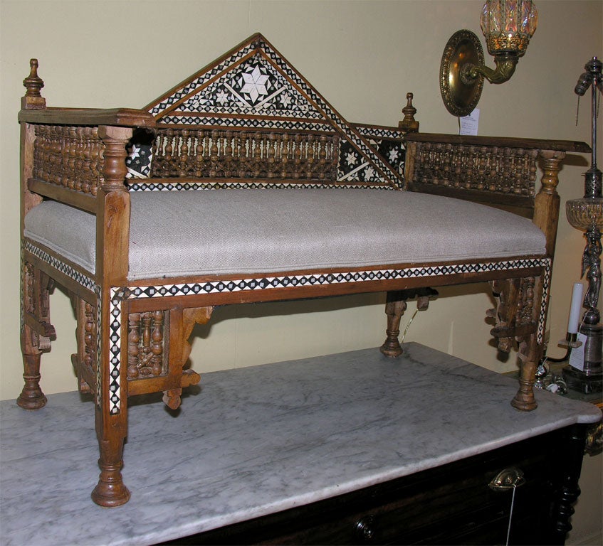 A Moroccan bench with bone and ebonized wood inlay, bobbin turned inserts, pediment backrest on newly upholstered seat resting on bun feet.  Unadorned back.