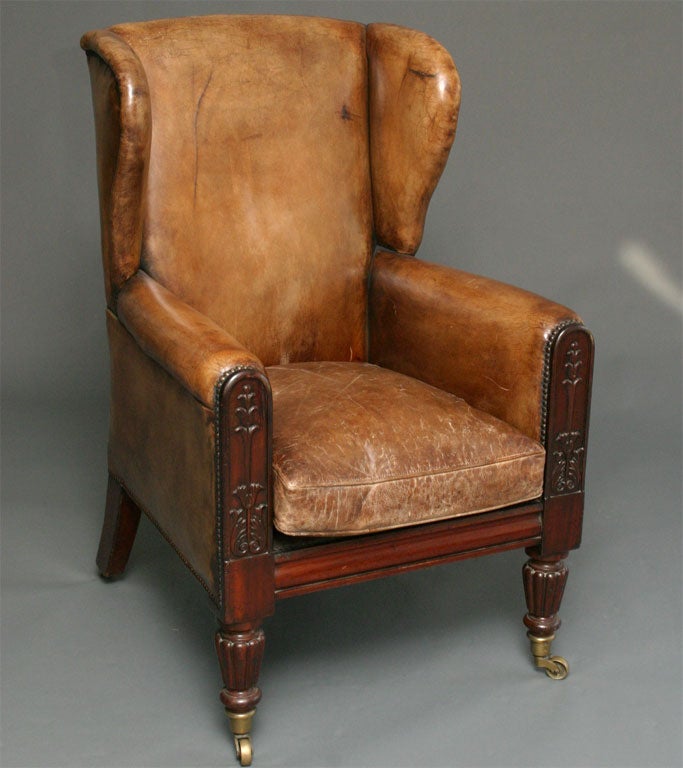 Victorian mahogany upholstered leather wing chair with loose padded cushion seat, antiqued nail heads, the arm supports carved with stylized foliage on reeded and turned legs with brass casters.