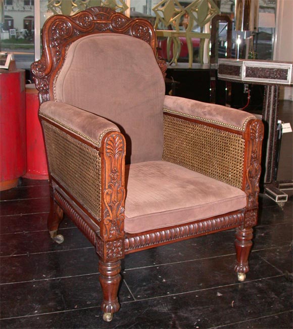 AN ANGLO-INDIAN GEORGE IV CANED PADOUK BERGERE THE FRAME CARVED IN LOW RELIEF WITH BOLD SCROLLWORK AND FLOWERHEADS WITH CONFORMING DESIGN ON THE REVERSE.   BEADED SEAT RAIL AND BRASS CAPS AND CASTERS  CANED SIDES AS WELL