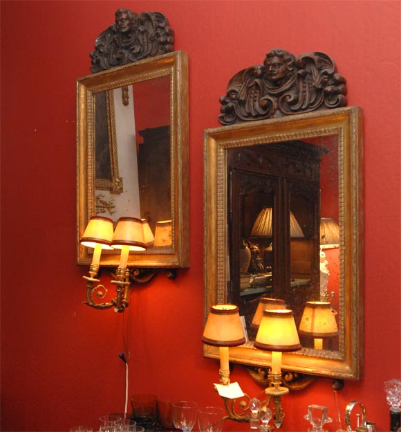 Each topped with a 17th century carved oak chapiteau of a male face    with scrolls and foliage, atop a 19th century gilt wood framed mirror, and two gilt brass scrolled arms terminating in candle sockets.