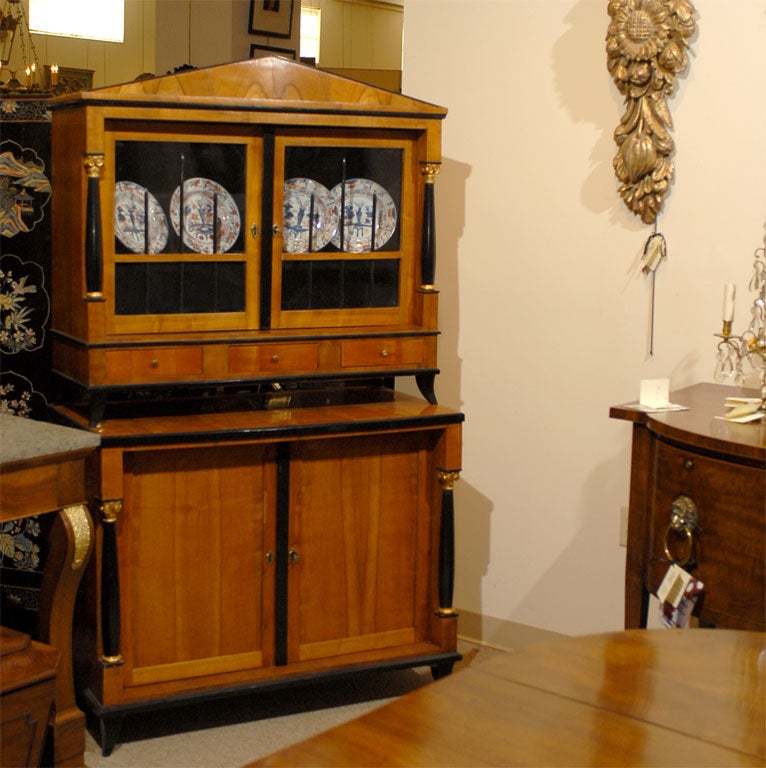 19th century German Biedermeier vitrine on cabinet base in fruitwood with ebonized and gilt accents. The display case with architectural pediment, fronted by glass panels with flanking ebonized columns and three drawers rests atop the lower cabinet