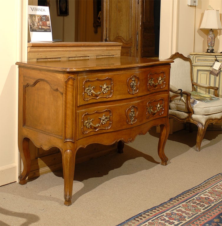 A Louis XV Period Serpentine Commode in Walnut with two sliding drawers featuring gilt-bronze fixtures, and mounted atop a carved shaped apron resting on cabriole legs.