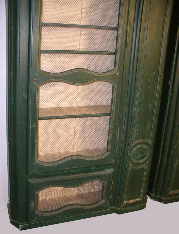 A pair of tall, green Louis XV style painted bookcases each having a large door fitted with handmade wire grill and enclosing adjustable book shelves, at the base of each cabinet is a smaller similarly fitted door revealing additional storage space.