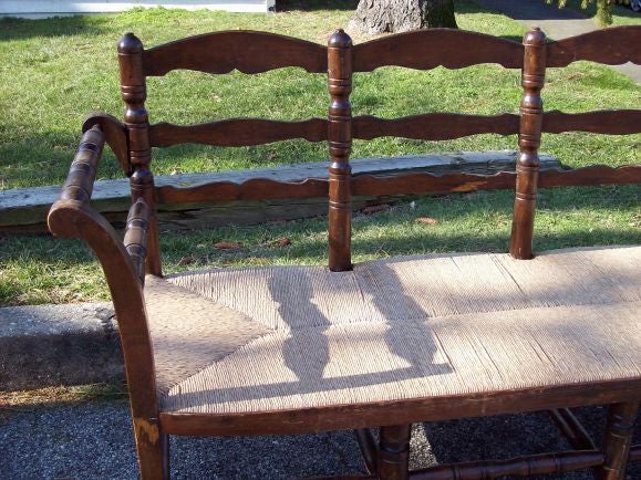 Antique rush seat bench from France. Seats four comfortably. The lovely curve of the arms and the beautifully turned legs and supports give this bench great charm. The wood has nice age and patina. Seat height is 15 1/2