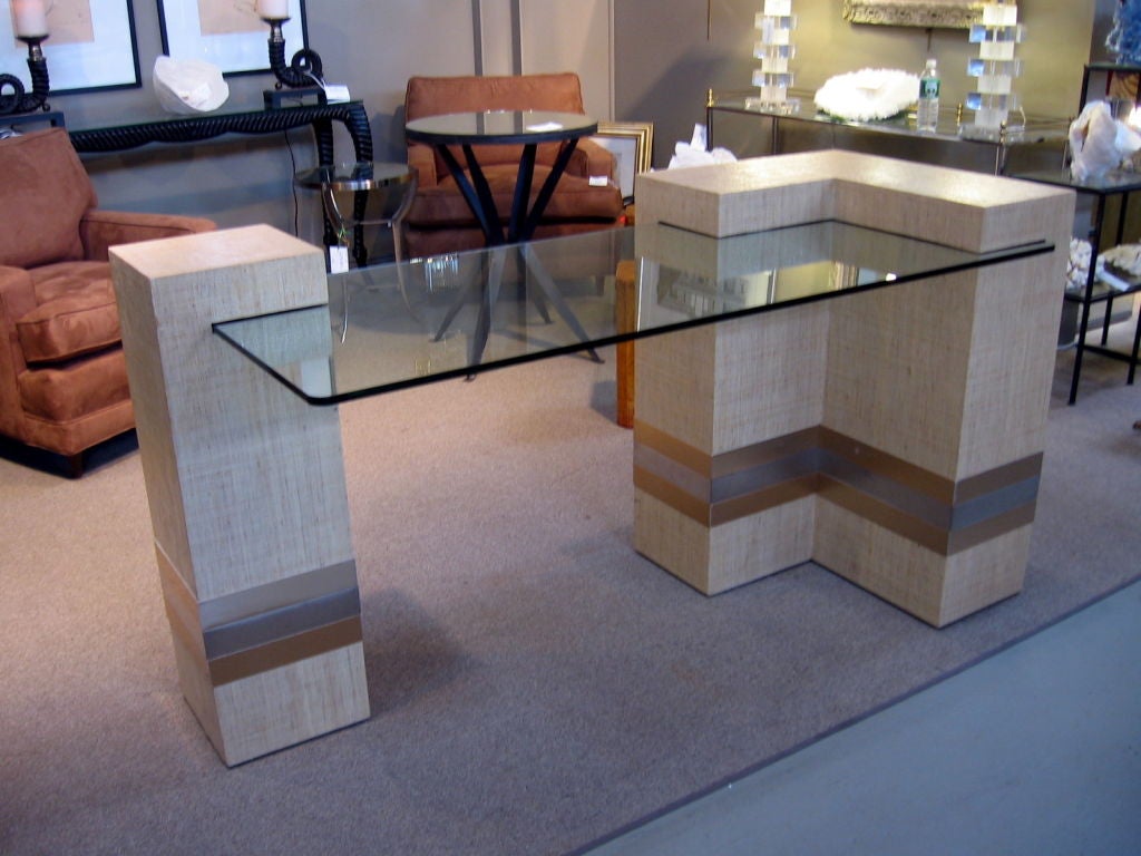 Asymmetrical writing table in the style of Karl Springer.  Glass top fits into asymmetrical  blocks that are covered in linen fabric that has been lacquered for durability.  There are three alternating bands of brass and silver near bottom.<br