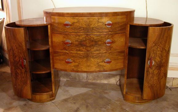 A chic and richly-patinated French art deco oval-form three-drawer sideboard flanked by cylindrical pedestals with hinged doors; of well-figured burl-walnut veneer and fitted with bakelite handles and aluminum backplates