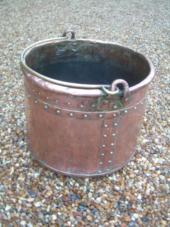 An English 19th century copper pot with hand rivetted seams and bottom, incorporating several old rivetted reinforcements, with a brass carrying handle.  Suitable for wood or kindling next to a fireplace.