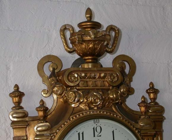 This baroque style cartel clock case has an eight day movement that strikes the hour and mid hour on a bell. The wooden case is gilded (with minor imperfections)and offers a variety of decorative features; flowers, ribbons, swags, scrolls, leaves