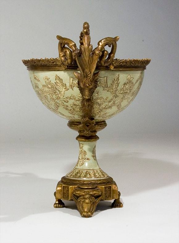 The brass ram’s head with stylized scolled horns of acanthus leaves attached to a brass crenalated edge. The bowl exterior and interior is a celadon ground enamel with verdant foliate patterns; raised on an elegant pedestal with brass footed raised