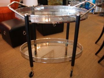 Rolling oval bar cart made of nickel silver with ebonized legs. Two tiered glass shelves with two handles.