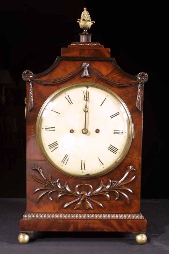 Regency mahogany bracket clock enriched with beautifully carved details. The shaped and stepped pediment surmounted with a brass ball finial. The sides with brass shell and ring handles above rectangular lozenge-pierced sound frets. The front with
