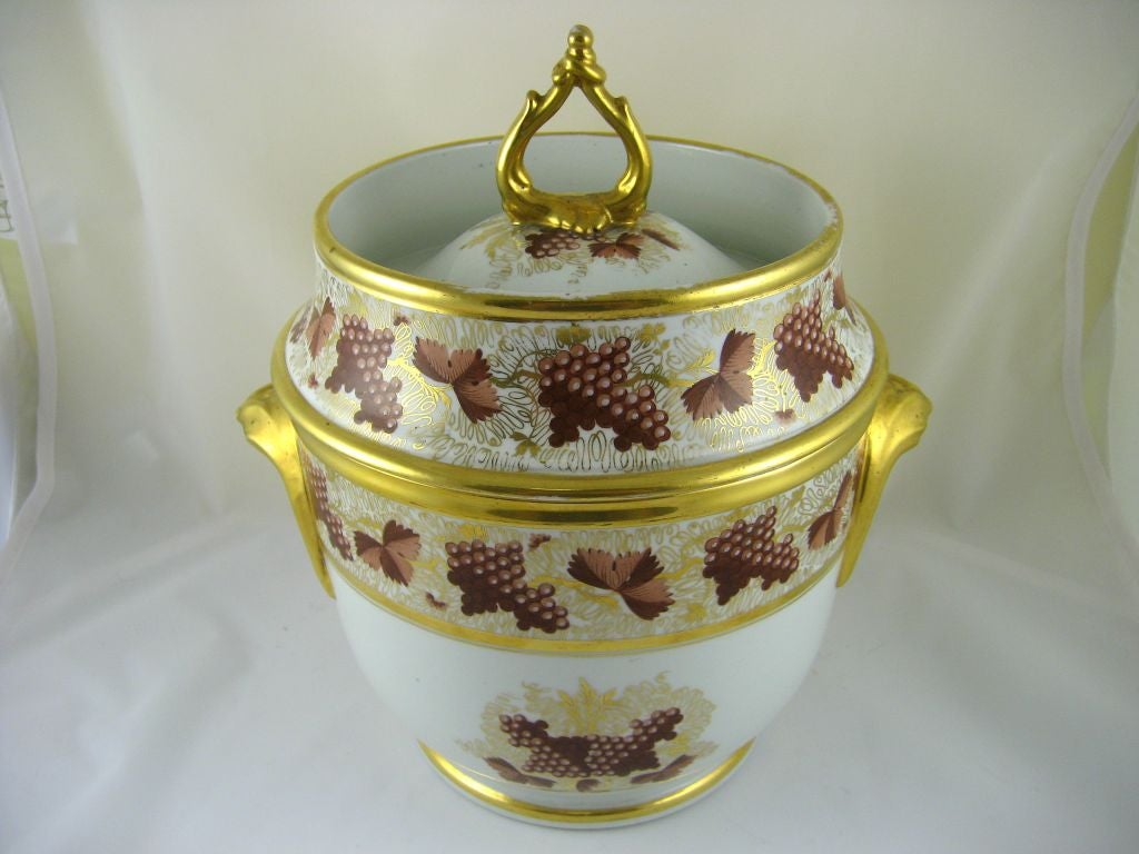 A Coalport Fruit Cooler and Cover, with intertwining vine finial atop a domed, recessed lid, and cusp handles on either side of the body. Painted in a mono-chromatic pattern simulating grapevines, with intricate gilt details designed in to emulate
