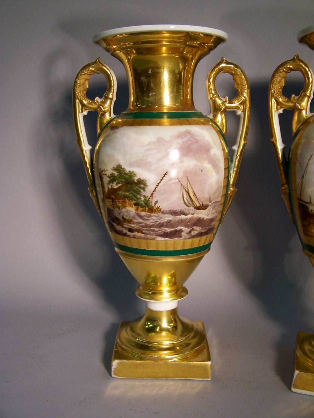 A fine pair of Paris porcelain vases on pedestal bases, featuring landscape scenes within acid-etched gilt borders. The body of each vase with a green ground, above and below with gilt ground. Each vase flanked by scrolled handles in gilt, with a